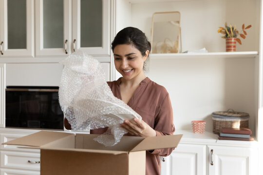 Smiling Indian woman unpacking fragile parcel, unwrapping plates, happy satisfied customer received online store order, buying kitchenware tableware in internet, happy renter unboxing belongings