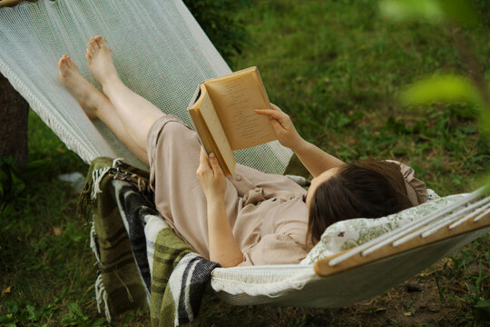 In a serene backyard, a woman finds escape in a book, swinging in a hammock. An embodiment of slow living and harmony, away from urban rush
