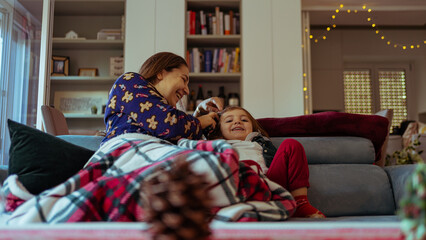 young single mother apprehensive combs her daughter on the sofa at home during winter vacation,...