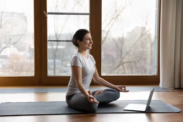 Fotobehang Lotusbloem Smiling Indian woman wearing grey sportswear meditating, using laptop, practicing yoga online, sitting on mat at home, happy mindful young female with closed eyes training, sitting in lotus pose  © fizkes