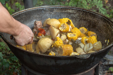 cook uses a slotted spoon to stir meat, corn on the cob, potatoes and vegetables in a cast-iron cauldron on an outdoor wood-burning stove. Street food