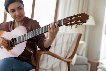 Close up Indian woman playing acoustic guitar, practicing chords, sitting in comfortable chair at...