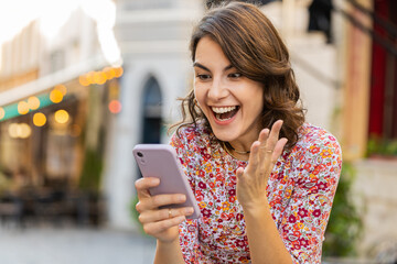 Portrait of woman use mobile smartphone celebrating win good message news lottery jackpot victory giveaway online play game outdoors. Happy girl tourist standing in urban city street. Town lifestyles - obrazy, fototapety, plakaty