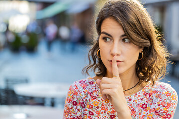 Shh be quiet please. Caucasian young woman presses index finger to lips makes silence hush gesture sign do not tells gossip secret rumors outdoors. Girl standing in urban city street. Town lifestyles - obrazy, fototapety, plakaty