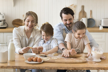 Fototapeta premium Young couple spend time with children in modern kitchen, parents teach little children to cook, prepare dough sit at dining table, have fun, make sweet homemade cookies. Family spend pastime together