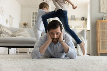 Young father exhausted with naughty active siblings, lies on floor in living room cover ears from...