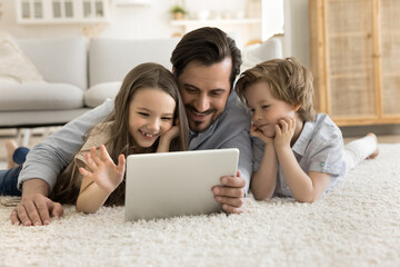 Smiling dad spend time with little kids at home, friendly family lying together on floor in living room use digital tablet, choose funny videos, chat remotely with relatives, make on-line purchases