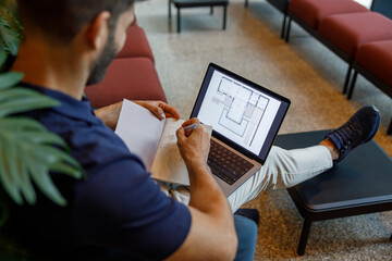 Close up of male architect is making notes in note pad during work on laptop in coworking space