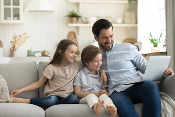 Happy handsome young father sits on sofa enjoy weekend time with cute little 6s children, hold laptop watch funny video, online TV family channel, doing e-shopping, laugh feels happy, use modern tech