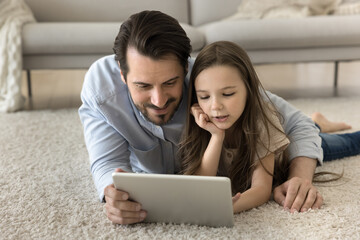 Smiling father and daughter choose funny movie on digital tablet lying on floor in living room, dad show new modern app to daughter, watch educational video, enjoy friendly relations, rest together