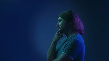 Side view of a mustachioed man with a headband in the studio on a blue background in pink and green neon light. A man is thinking intently about something, putting his hand to his chin.