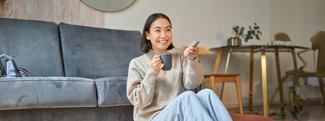 Portrait of young korean woman watching television, holding remote and looking amazed at tv screen,...