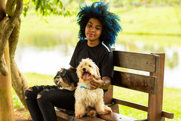 Jovem mulher negra com cabelos pintados de azul, com seus dois cachorros em um banco do parque.