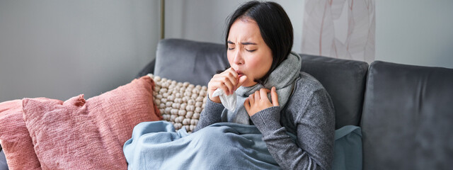 Asian woman coughing, feeling sick, sitting under blanket with scard in cold apartment, saving on heating this winter, catching flu - obrazy, fototapety, plakaty