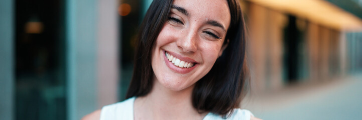 Fototapeta premium Close-up of young woman with freckles and dark loose hair and long eyelashes wearing white top looking straight at the camera. Beautiful girl on modern city background