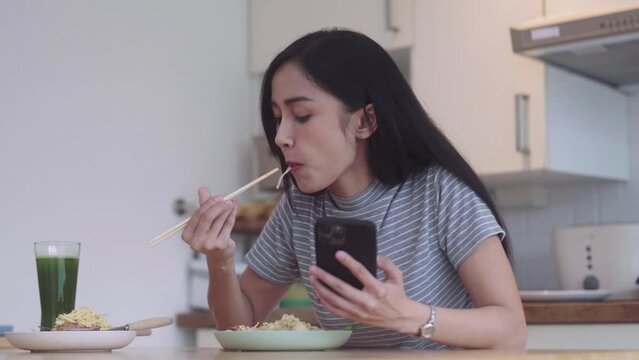 joyful nice Asian Woman having asian noodles food pad thai holding chopsticks for lunch during lunch break and typing in her smartphone or browsing social networks on the table in kitchen at home