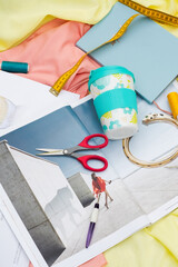 Fototapeta premium sharp red scissors, violet spreader, blue notebook and yellow measuring tape and laying cup for tea on open magazine. Pink and yellow soft fabric. Sewing set. Girl on pages of magazine 