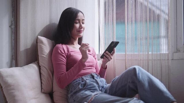 Cheerful beautiful pretty young Asian woman sitting on a windowsill at home and texting on her phone communication and using app on Internet near window at home.