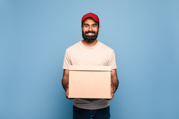 Delivery service concept. Cheerful indian mailman holding cardboard box and smiling at camera, blue...