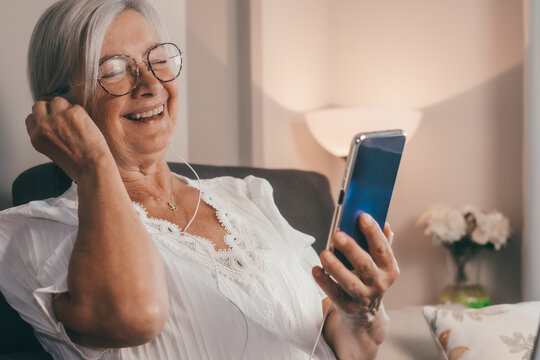 Happy Laughing Senior Retired Woman Sitting On Sofa Wearing Earphones, Using Mobile Phone Technology, Enjoying Carefree Moments Communication Chat Video Call With Family Or Friends