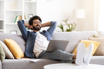Relaxed Young Indian Man With Laptop Sitting On Couch At Home Interior