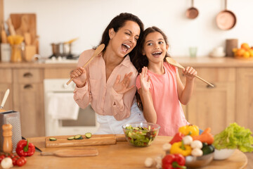 Mom And Daughter Singing With Spoons Preparing Dinner In Kitchen - obrazy, fototapety, plakaty