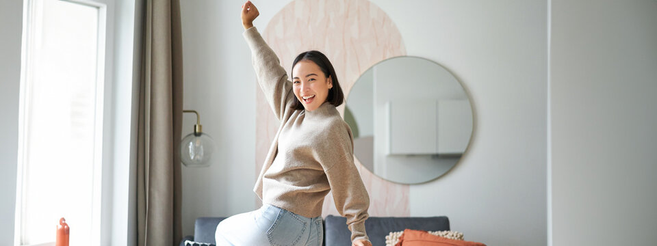 Cute Excited Asian Woman Dancing, Raising Hands Up In Triumph, Celebrating And Singing, Standing At Home In Warm Jamper