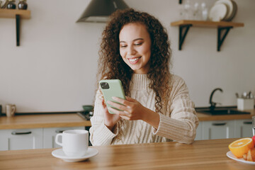 Young smiling happy housewife woman wearing casual clothes sweater hold use mobile cell phone drink...