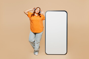 Full body young chubby overweight woman wear orange shirt casual clothes big huge blank screen area mobile cell phone listen to music in headphones isolated on plain beige background studio portrait.