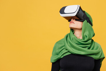 Young gambling arabian asian muslim woman wear green hijab abaya black clothes watching in vr headset pc gadget isolated on plain yellow background. People uae middle eastern islam religious concept.