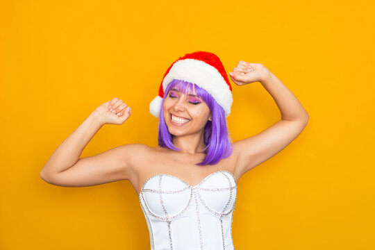 A young happy smiling caucasian woman with violet hair in white festive corset, red Santa Claus hat dancing with her hands up isolated on a color yellow background. Christmas and New Year concept