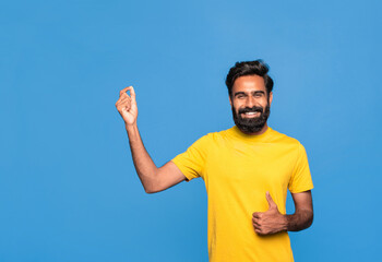 Excited indian man holding something invisible in hand and showing thumbs up gesture, standing over blue background