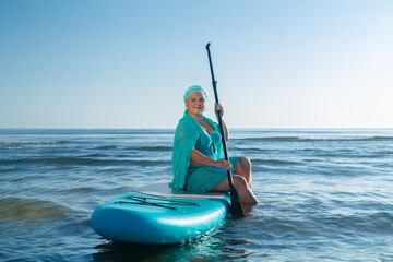 A woman in a turquoise swimsuit with a skirt and a scarf on her head sits on a SUP board near the...