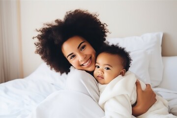 Young African American mother holding her healthy sleeping newborn baby in bed at home. Mother's day concept. Loving mom looking at camera and smiling.
