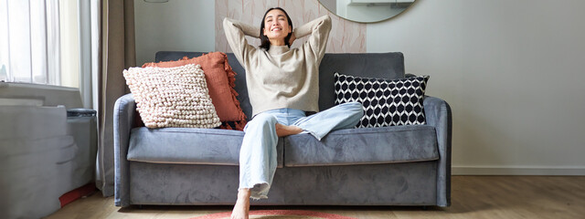 Portrait of carefree asian woman enjoying day-off, sitting on sofa and smiling pleased, relaxing in house on weekends - obrazy, fototapety, plakaty