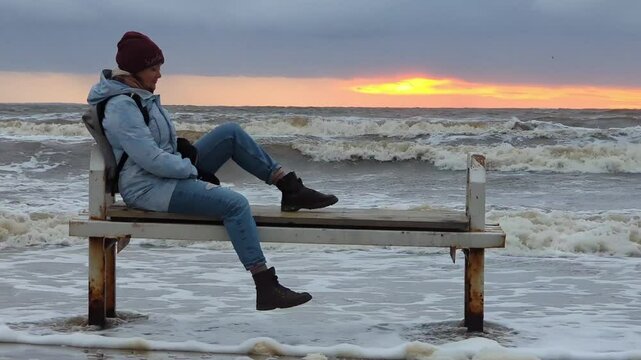 Lonely girl sitting on a bench by the sea in a winter storm. High quality FullHD footage