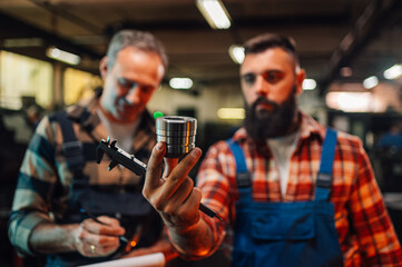 Focus frame of a hand holding a caliper and a metal piece, two people blurred in background.