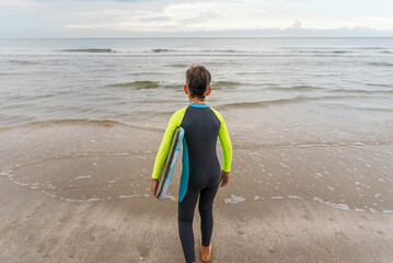 Young boy in a wet suit at the beach walking into the water while carrying his boogie board