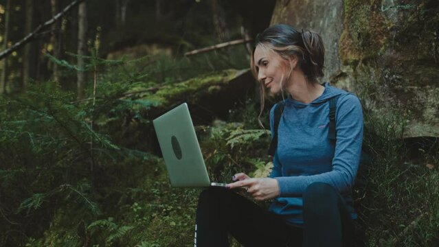 Woman work on laptop sitting near moss stone in sunset forest. Tourist girl typing on notebook, rest stop in hiking route. Outdoor lifestyle travel summer holiday vacation. Camera rise up, close up 4K