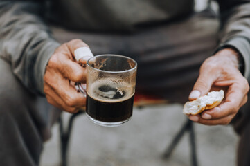 Old, elderly, homeless, poor male worker holds in his hands a glass cup of coffee, croissant, pastry during lunch, snack. Close up food photography, poverty concept.