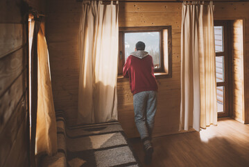 A man in cozy home clothes stands in a cozy room in a wooden cottage and admires the view of the...