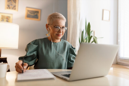 Indoor image of attractive stylish elegant woman in eyeglasses watching video tutorial in wired earphones, noting down main things in her textbook, sitting near window at table desk at home