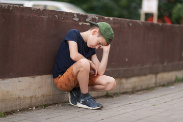 A teenage boy is squatting leaning against a concrete wall on the street. Upset and sad. Bullying....