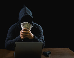 Hacker spy man one person black hoodie sitting on table hand holding money look computer laptop...