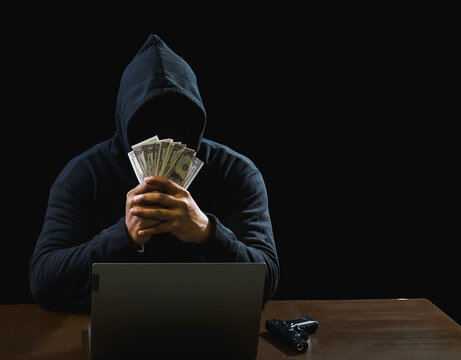 Hacker spy man one person black hoodie sitting on table hand holding money look computer laptop used login password attack security to circulate data digital internet network system, dark background.