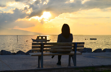  A woman sitting on a bench near a sea and looking at cloudy sunset