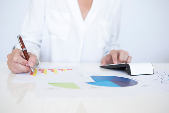 Detail of a middle-aged woman in a white shirt sitting at a white table, working with documents with graphs and financial statistics holding a pen in her right hand.