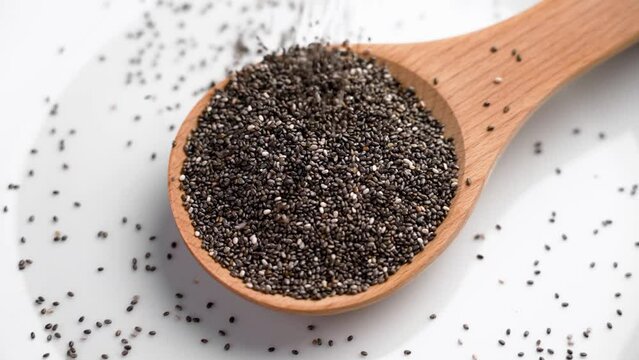 Raw nourishing chia seeds in kitchen wooden spoon on white ceramic surface. Falling dried particles in slow motion. Rotation