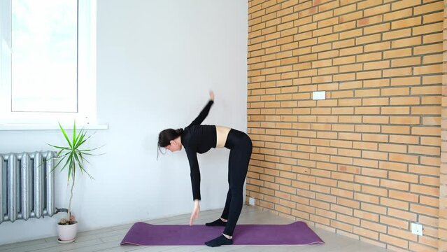 Beautiful sporty 30s woman is doing windmill exercise at home, having an active morning, strengthening the core and spine muscles, fitness and health concept. Healthy body practicing. Black clothing.