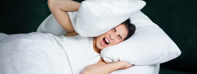 Sad girl with insomnia. Young asian woman lying in bed, cover her ears with pillow, cant sleep, partner snors, loud noise at night disturbing her - obrazy, fototapety, plakaty